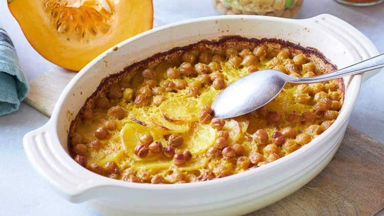 Ein Auflauf mit Kichererbsen und Kartoffelscheiben in einer weißen Ofenform, daneben ein halber Kürbis und ein Löffel.