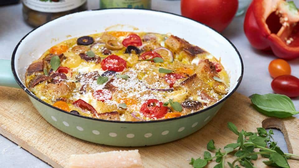 Eine grüne Auflaufform mit Punkten enthält eine bunte Gemüse-Frittata, dekoriert mit frischen Kräutern, auf einem Holzbrett.