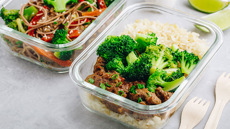 Zwei Glasbehälter mit gekochtem Essen, vorn Brokkoli, Reis und Hackfleisch, hinten Gemüse mit Nudeln, Holzgabel rechts.