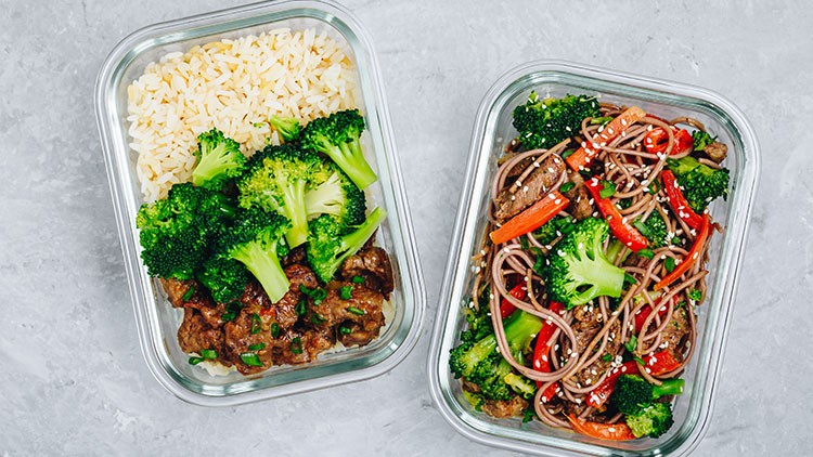 Zwei Glasbehälter mit Reis, Brokkoli und Rindfleisch links sowie Nudeln, Brokkoli und Paprika rechts auf grauem Untergrund.
