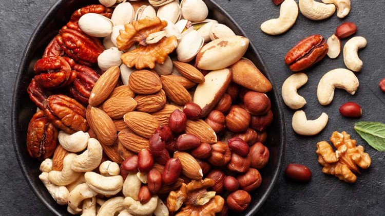 Schale mit verschiedenen Nüssen wie Mandeln, Cashews und Walnüssen, teils auch neben der Schale auf dunklem Untergrund.