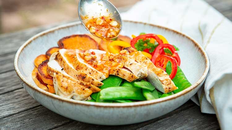 Eine Schüssel mit Hähnchenstreifen, Süßkartoffelscheiben, Zuckerschoten und Paprika, darüber wird Sauce aus einem Löffel gegeben.
