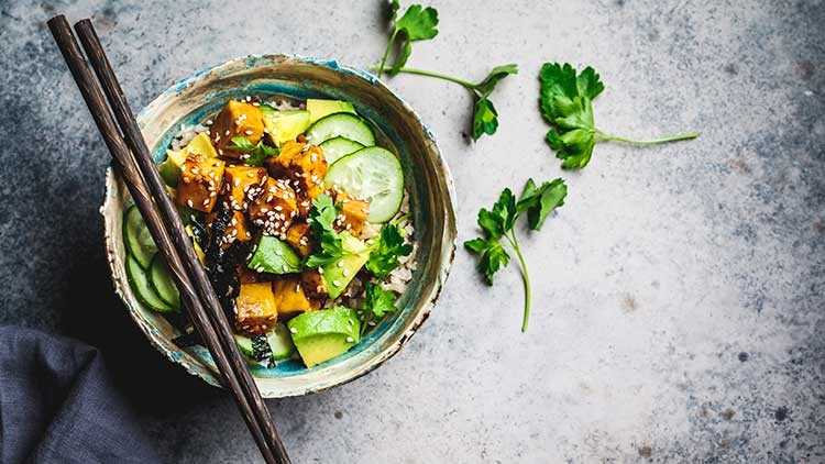 Bunte Schüssel mit Reis, Gurken, Avocado, würfeligem Tofu, Sesam und Kräutern, daneben zwei Holzstäbchen und Petersilie.
