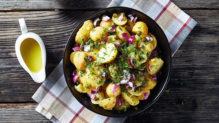 Schüssel mit Kartoffelsalat, garniert mit frischen Kräutern, roten Zwiebeln und Kapern, daneben eine weiße Sauciere mit Dressing.