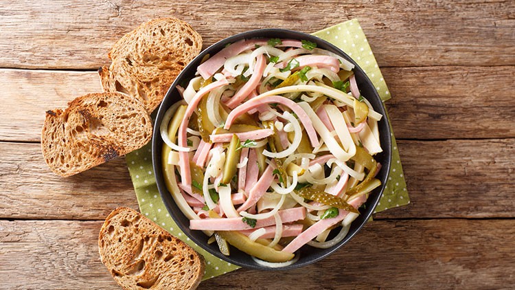 Eine Schüssel Wurstsalat mit Zwiebeln, Essiggurken und Kräutern steht auf einem Holzuntergrund mit gerösteten Brotscheiben.