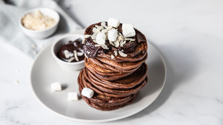 Ein Stapel Schokoladenpfannkuchen mit Marshmallows, Mandelsplittern und Schokosoße, daneben eine kleine Schüssel Schokosoße.