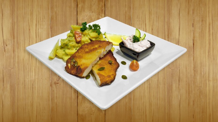 Panierter, goldbrauner Schnitzelersatz mit Kartoffelsalat, Zitronenspalte, Kürbiskernen und Dip in schwarzem Schälchen auf weißem Teller.