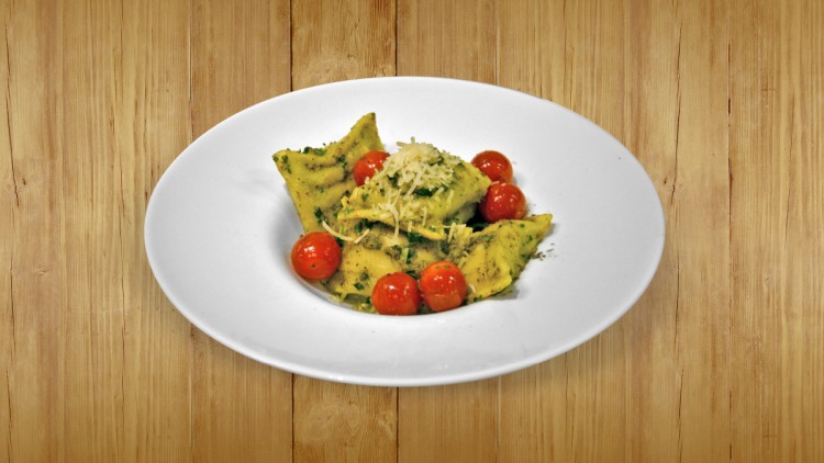 Gefüllte Ravioli mit Pesto, kleinen Tomaten und geriebenem Käse auf weißem Teller vor hellem Holzuntergrund.