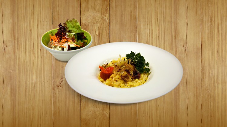 Ein Teller mit Käsespätzle, Röstzwiebeln, einer roten Tomate und Petersilie, daneben eine Schüssel mit gemischtem Salat.