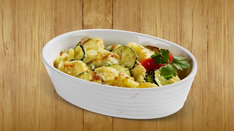 Weiße Auflaufform mit gebackenem Zucchini-Käse-Auflauf, garniert mit einem roten Tomatenstück und einem Zweig frischer Petersilie.