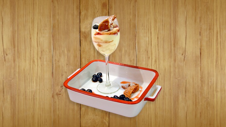 Ein mit Creme, Blaubeeren und Gebäck gefülltes Weinglas steht in einer weißen Backform mit rotem Rand auf Holzuntergrund.
