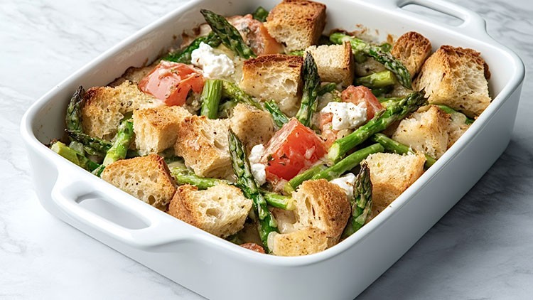 Weiße Auflaufform mit gerösteten Brotwürfeln, grünem Spargel, Tomatenstücken und zerbröseltem Käse auf hellem Untergrund.