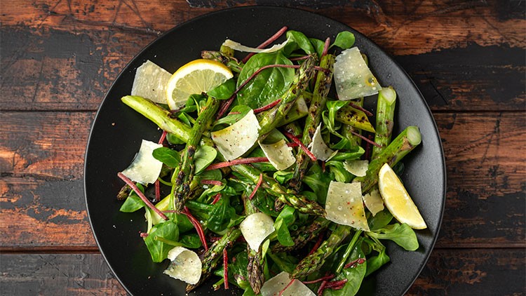 Grüner Salat mit gegrilltem Spargel, Parmesanflocken, Zitronenscheiben und roten Blättern auf einem schwarzen Teller.