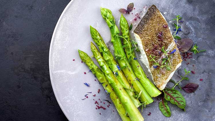 Gebratener Fisch mit knuspriger Haut, grünem Spargel und Kräuterdekoration auf einem hellen Teller, darunter essbare Blüten.