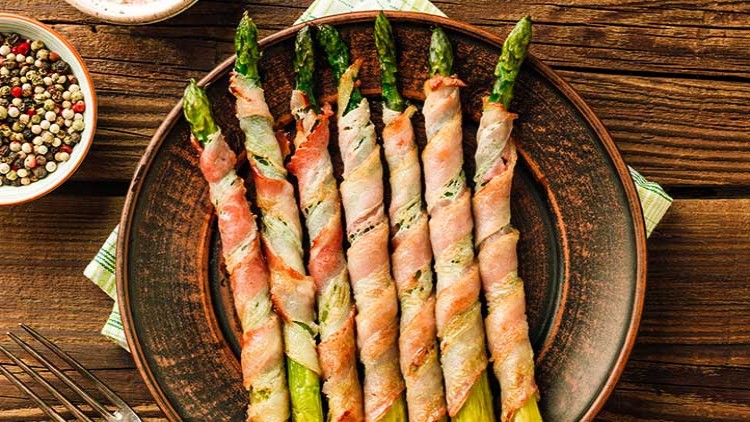 Grüner Spargel, umwickelt mit knusprigem Schinken, liegt auf einem braunen Tellerrand, mit Pfefferkörnern in einer Schale daneben.