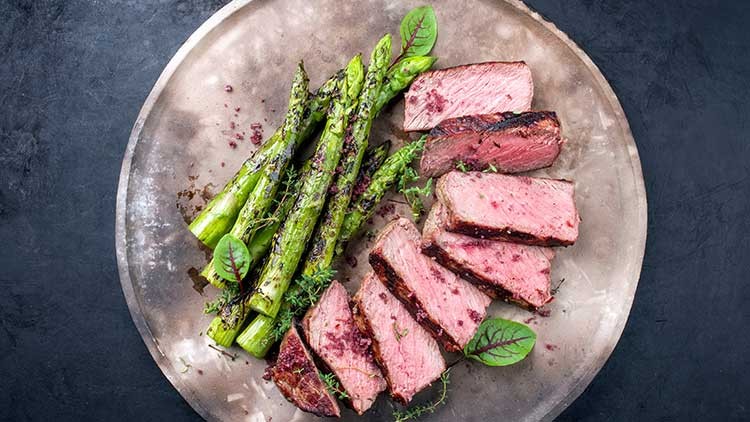 Gegrillter grüner Spargel und in Scheiben geschnittenes rosa gebratenes Fleisch auf einem Teller mit frischen Kräutern.
