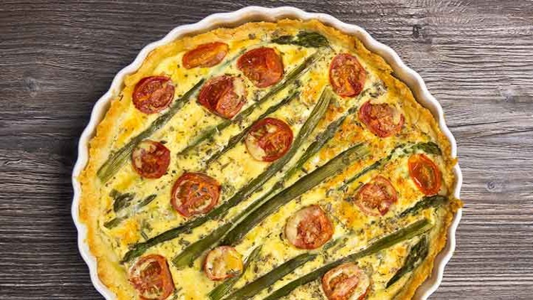 Herzhafte Quiche mit grünem Spargel und Tomatenscheiben in einer runden weißen Backform auf dunklem Holzuntergrund.
