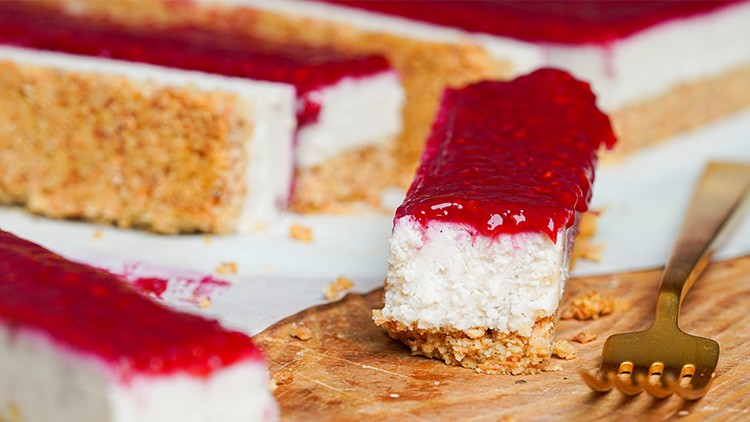 Ein Stück Käsekuchen mit roter Fruchtschicht und knusprigem Boden liegt auf einem Holzbrett neben einer goldenen Gabel.