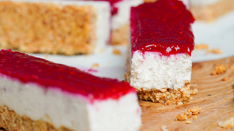 Riegel aus Haferkeksboden mit heller Creme und roter Fruchtsoße darauf, auf einem Holzbrett und weißem Untergrund.