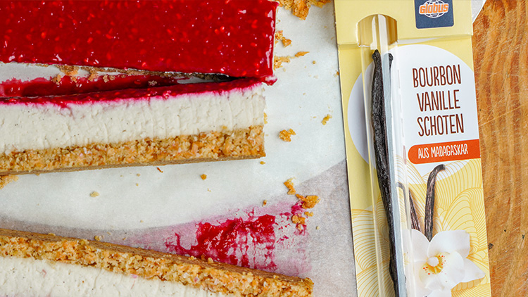 Stück Käsekuchen mit roter Fruchtschicht neben einer Verpackung von GLOBUS Bourbon-Vanilleschoten aus Madagaskar.