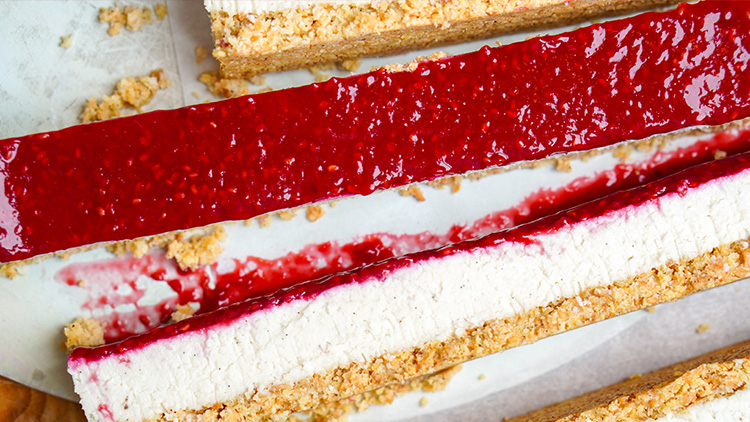 Rote Fruchtschicht auf cremigem Belag mit knusprigem Boden, umgeben von Krümeln auf einer weißen Unterlage.