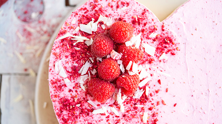 Herzförnige Torte mit rosa Creme, dekoriert mit frischen Himbeeren, roten Krümeln und weißen Schokoladensplittern.