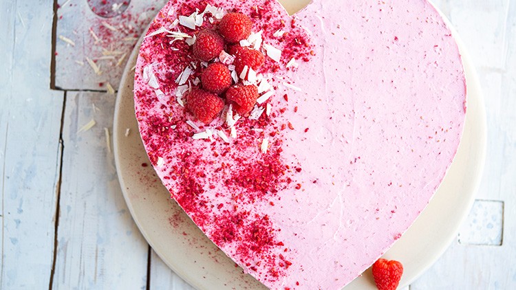Herzförnige Himbeertorte mit rosa Creme, Himbeeren, weißen Schokoraspeln und rotem Streudekor auf einem Teller.