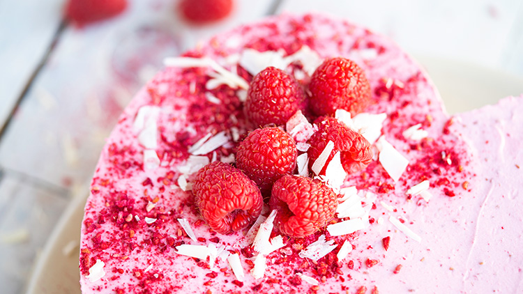 Rosa Kuchen in Herzform mit frischen Himbeeren, roten Streuseln und weißen Schokoladenraspeln dekoriert.