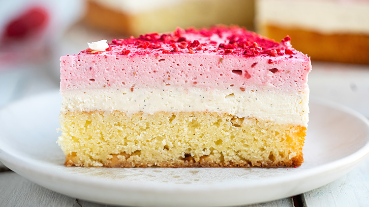 Ein Stück Kuchen mit hellem Boden, weißer und rosafarbener Creme, dekoriert mit roten Streuseln, auf einem weißen Teller.