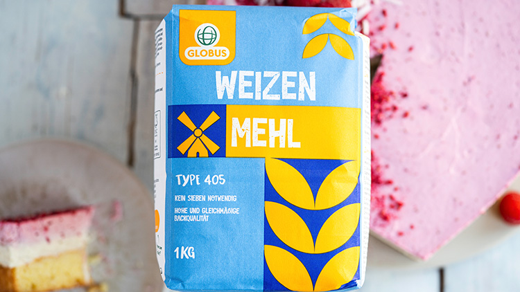 Eine 1-kg-Packung GLOBUS Weizenmehl Type 405 steht vor einem rosa Kuchen und einem Stück Kuchen auf einem Teller.