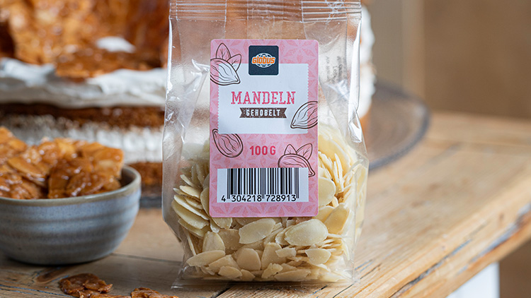 Eine Plastiktüte mit der Aufschrift „MANDLN GEMAHLEN, 100 G“ steht auf einem Holztisch, daneben liegt ein Schüssel.