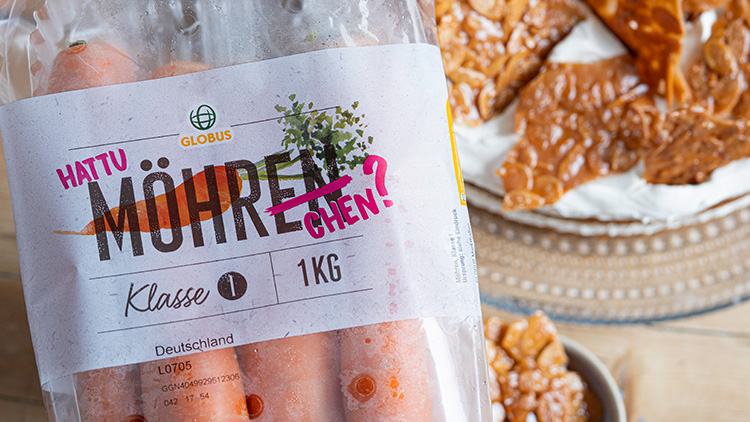 Karotten in einer durchsichtigen Verpackung mit der Aufschrift „Hattu Möhrchen?“, daneben Gebäck auf einem Teller.