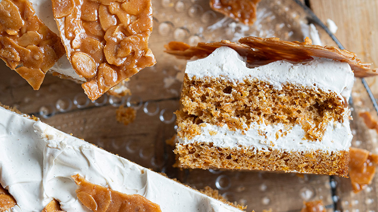 Ein Stück Lebkuchen mit weißer Creme und knusprigem Karamell auf einer gläsernen Platte, umgeben von weiteren Stücken.