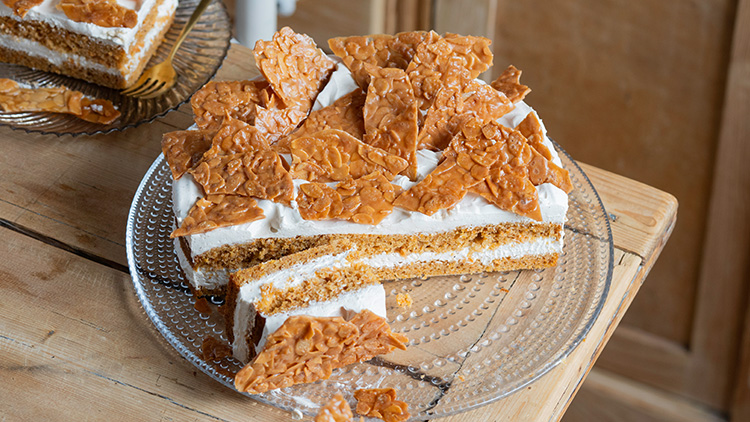 Runde Torte mit weißer Creme-Schicht, dekoriert mit goldbraunen Mandel-Krokant-Stücken auf einem Glasteller.