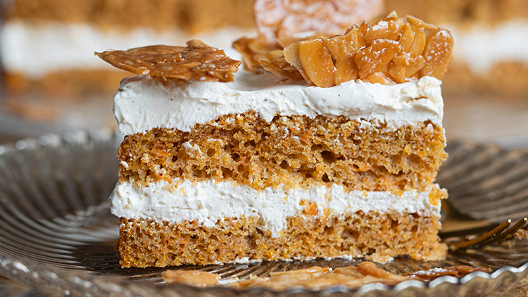 Ein Stück saftiger Kuchen mit heller Creme und Karamellstücken liegt auf einem gläsernen Teller, daneben eine goldene Gabel.