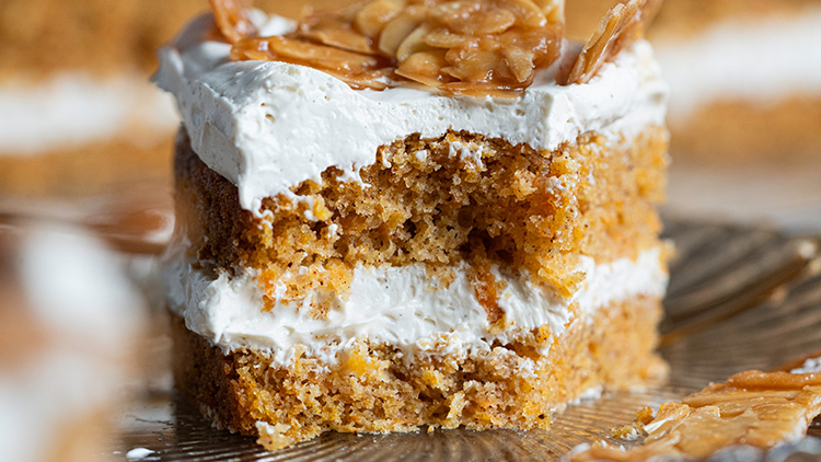 Ein Stück Kuchen mit heller Cremefüllung, oben mandelkaramellierte Dekoration, auf einem Glasteller.
