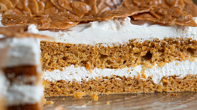 Ein angeschnittener Kuchen mit hellbraunen Böden, einer hellen Cremefüllung und knusprigen karamellisierten Mandelscheiben oben.