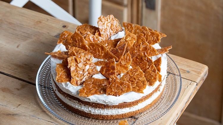 Torte auf Glasplatte mit weißer Creme und karamellbraunen Knusperstücken, auf Holztisch vor hellem Hintergrund.
