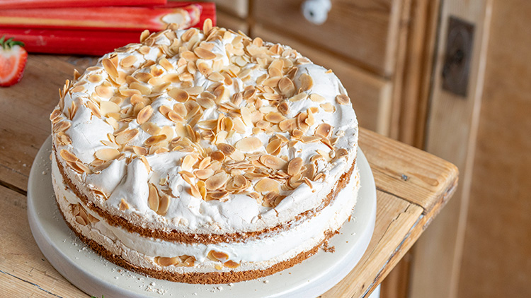 Runde Sahnetorte mit knusprigen Mandelblättchen auf einem weißen Teller, daneben rote Rhabarberstangen auf einem Holztisch.