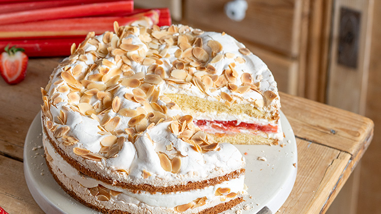 Rhabarber-Biskuit-Torte mit weißer Schlagsahne, gerösteten Mandelblättchen und einer sichtbaren fruchtigen Schicht auf Teller.