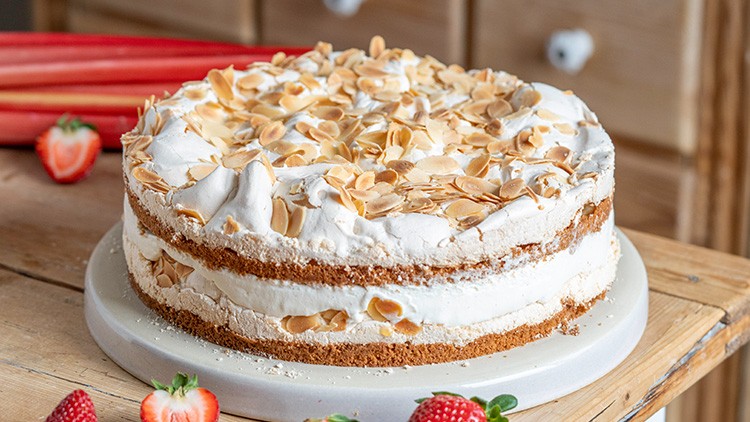 Runde Sahnetorte mit Baiser und gerösteten Mandelblättchen auf einem Teller, umgeben von Erdbeeren und Rhabarberstangen.