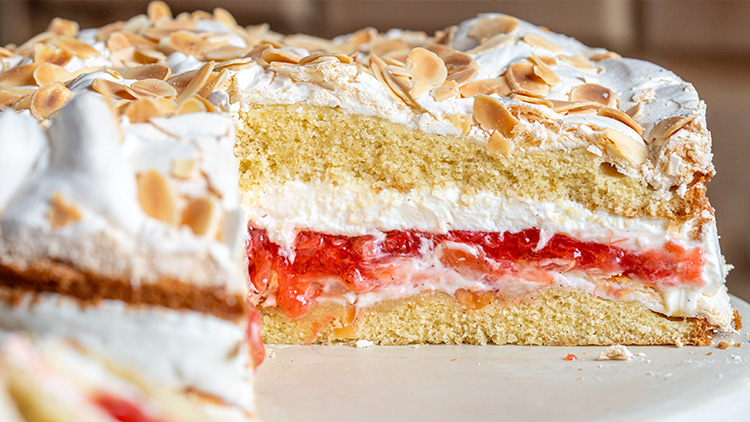 Eine aufgeschnittene Torte mit Schichten aus Biskuit, Sahne, roter Fruchtfüllung und gerösteten Mandelblättchen oben.
