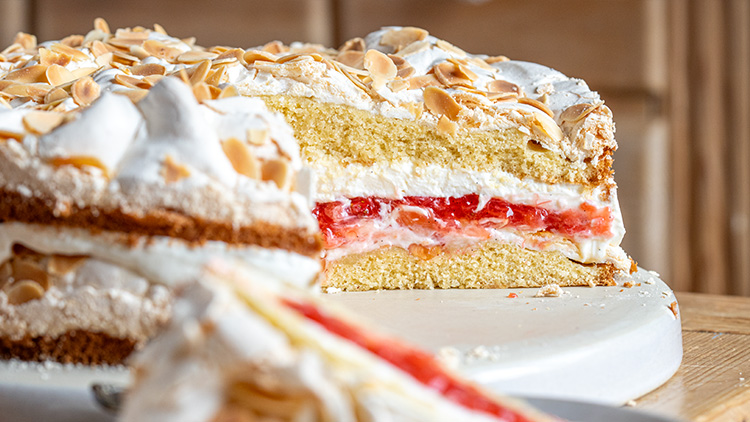 Eine Torte mit Schichten aus Biskuit, Sahne und rotem Kompott, dekoriert mit Mandelsplittern, auf einem weißen Teller.