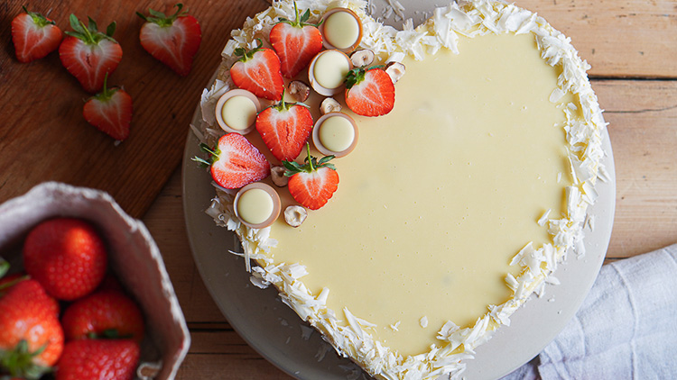 Herzförmige Torte mit weißer Glasur, dekoriert mit Erdbeeren, weißer Schokolade und Kokosraspeln auf einem Holztisch.
