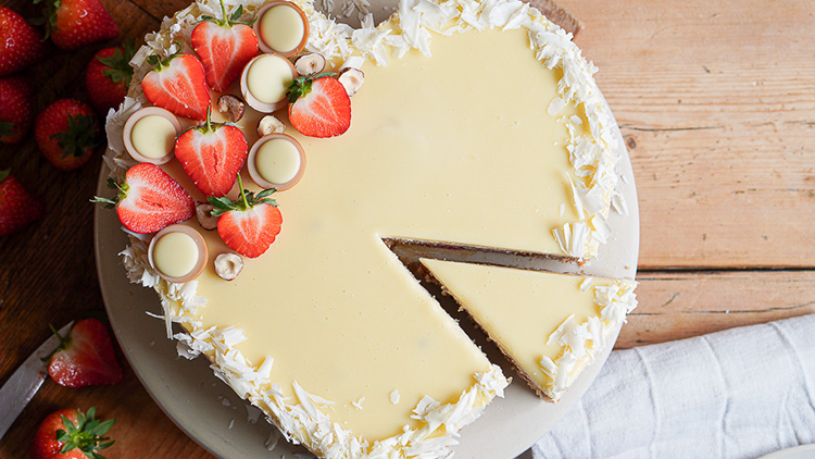 Rundherum mit weißer Schokolade verzierte Torte, belegt mit Erdbeeren, Haselnüssen und Pralinen, auf einem Holztisch.
