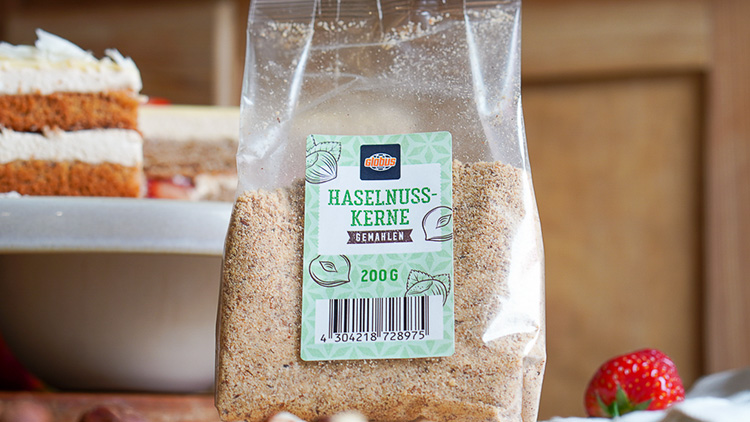 Eine Packung gemahlene Haselnusskerne von GLOBUS, 200 g, steht vor einem Kuchenstück auf einem Tisch mit Erdbeeren.