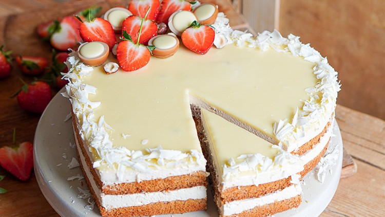 Eine herzförmige Torte mit heller Glasur, Schichten aus Creme und Teig, dekoriert mit Erdbeeren und weißen Schokoraspeln.