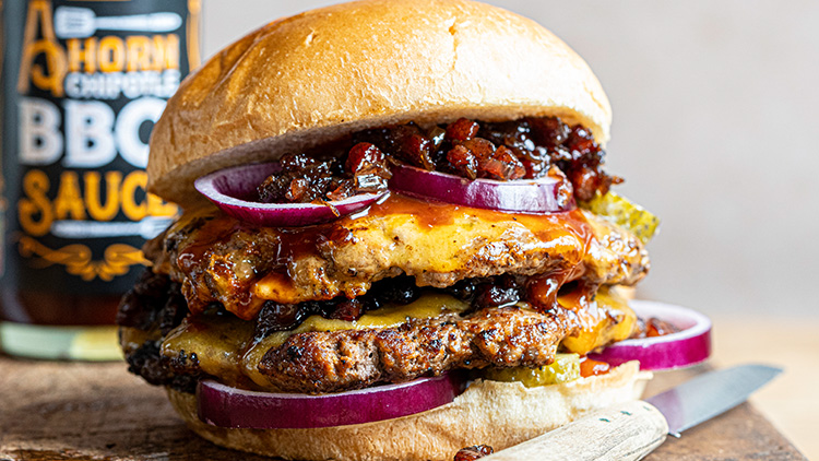 Ein Burger mit zwei Fleischpattys, Käse, roten Zwiebeln, Essiggurken, Barbecuesoße und Speck auf einem Brötchen.