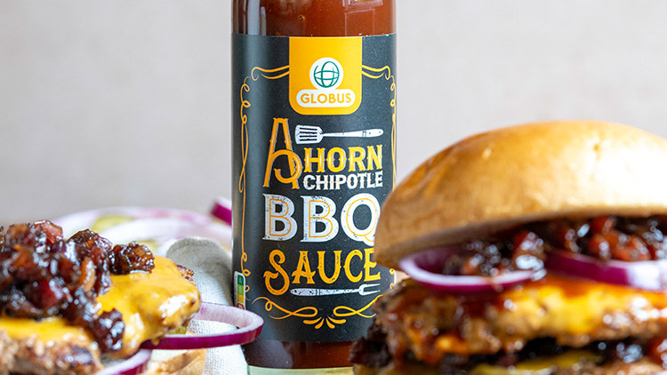 Ahorn Chipotle BBQ Sauce von GLOBUS mit gelbem Logo, im Vordergrund Hamburger mit Käse, roten Zwiebeln und dunkler Sauce.