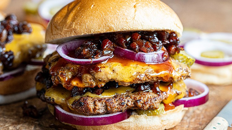 Burger mit zwei Fleisch-Patties, geschmolzenem Käse, roten Zwiebelringen, Gurkenscheiben und karamellisierten Zwiebeln im Brötchen.